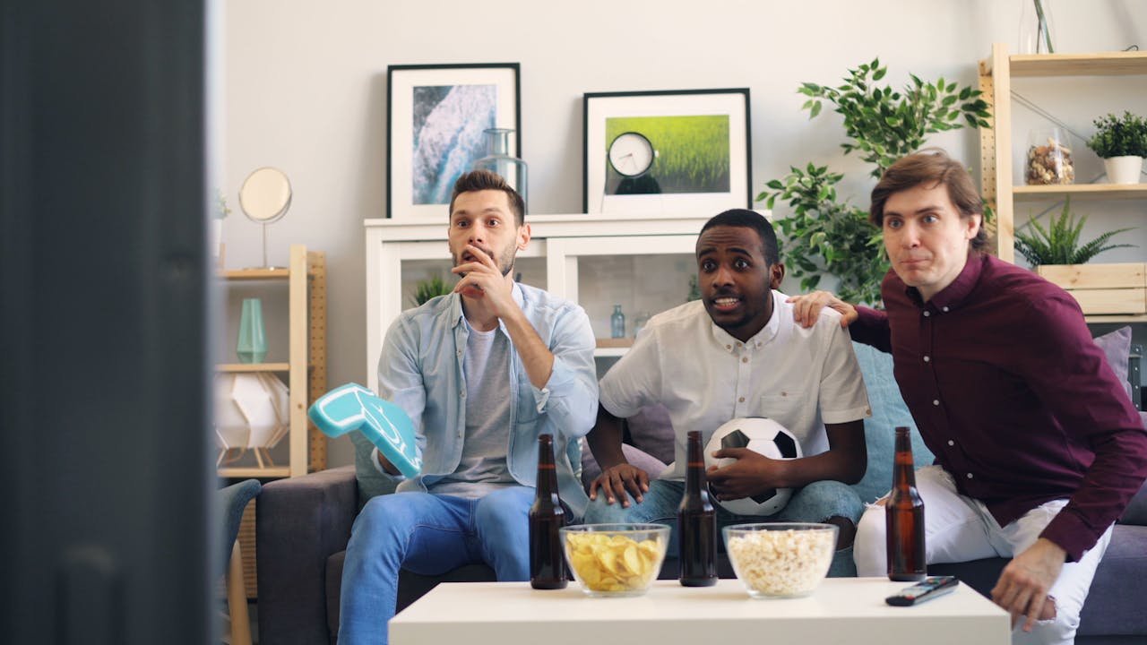 Three friends excitedly watch a soccer game at home, showing emotions and enjoying snacks.